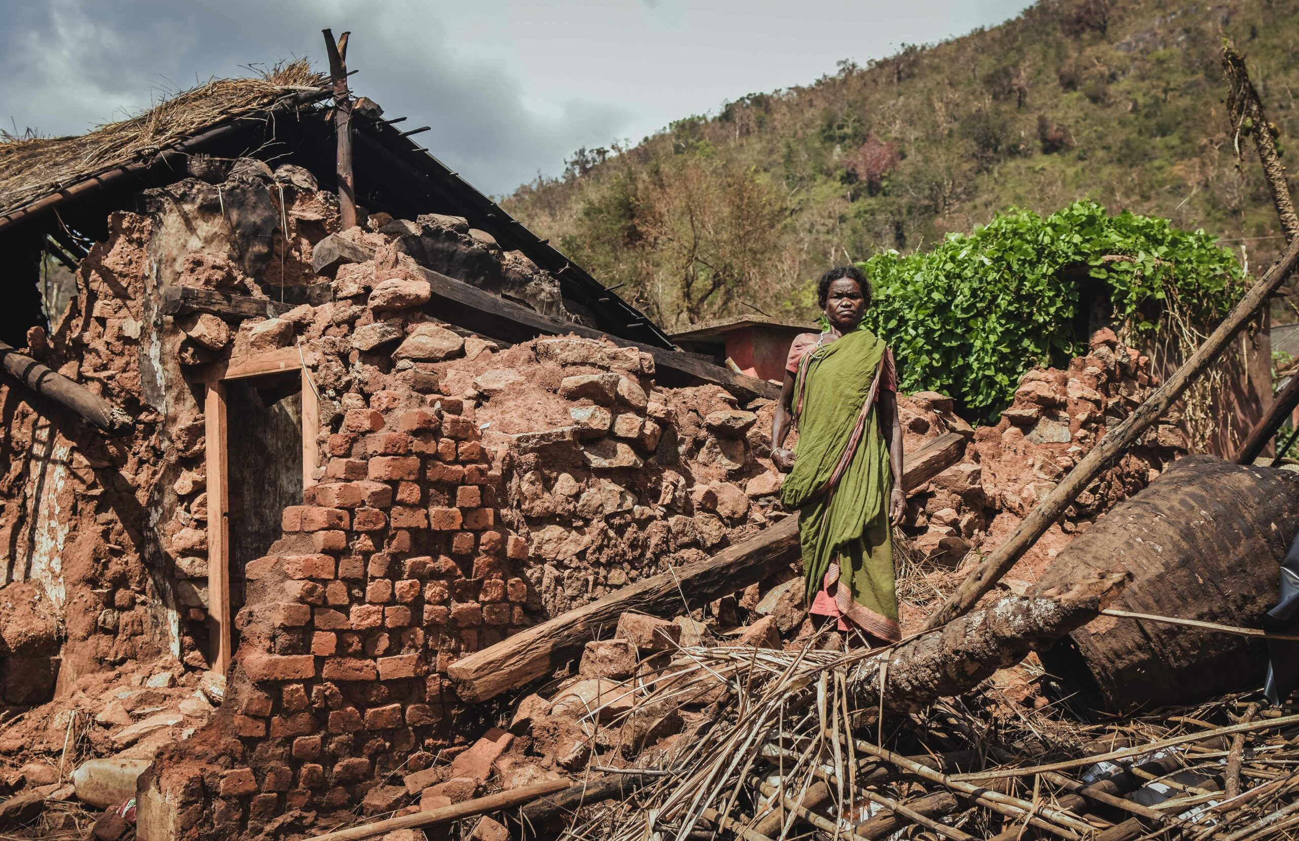 Our Areas Woman stands in front of a destroyed house after a disaster in rural India, depicting struggle.